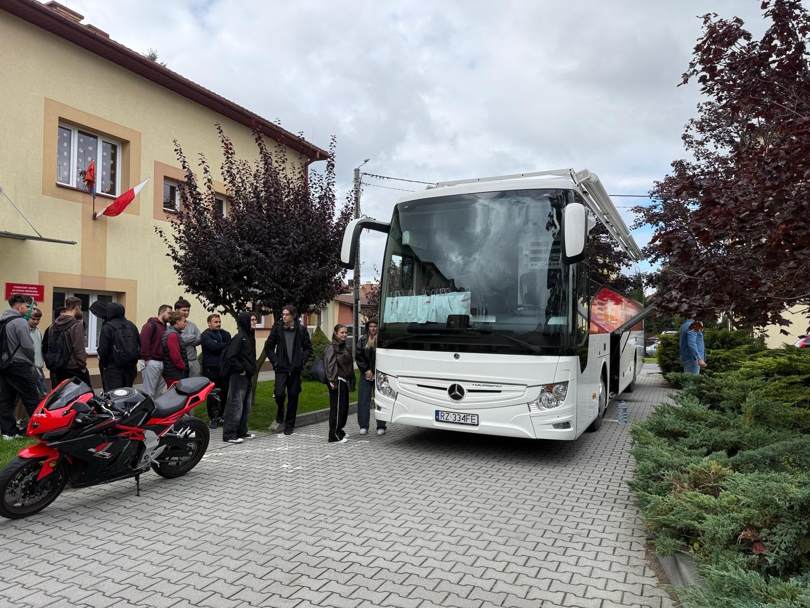 Przed budynkiem szkoły stoi mobilny punkt poboru krwi, obok czekają uczestnicy akcji.