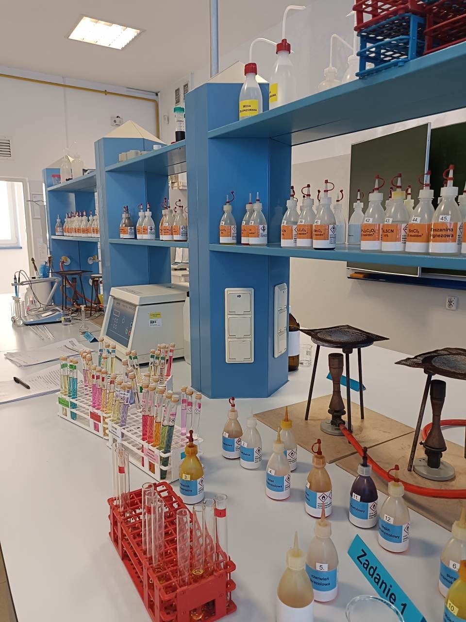 Stanowisko laboratoryjne z odczynnikami, probówkami i sprzętem do doświadczeń chemicznych.