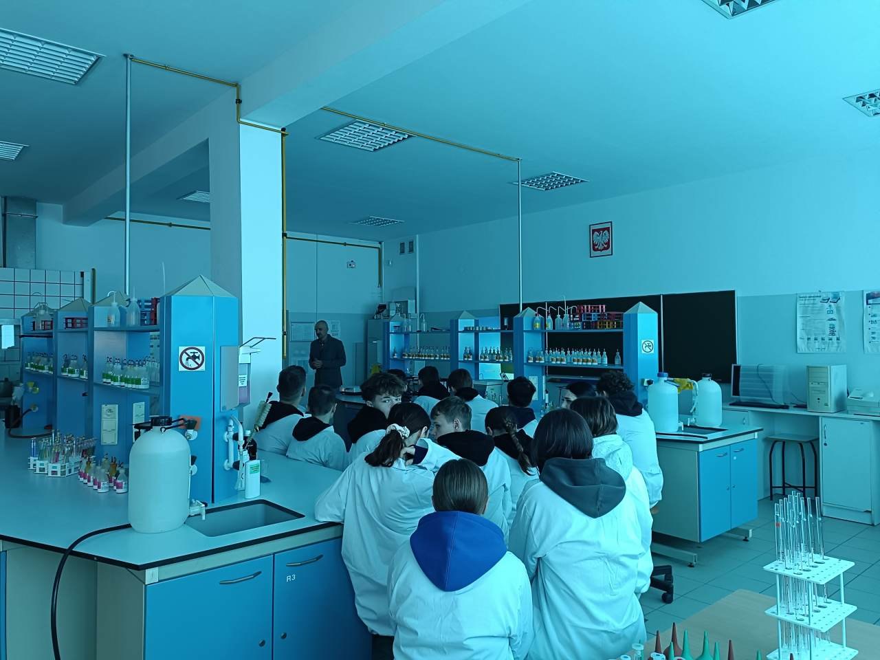 Uczniowie w fartuchach laboratoryjnych słuchają wprowadzenia do zajęć w pracowni chemicznej.