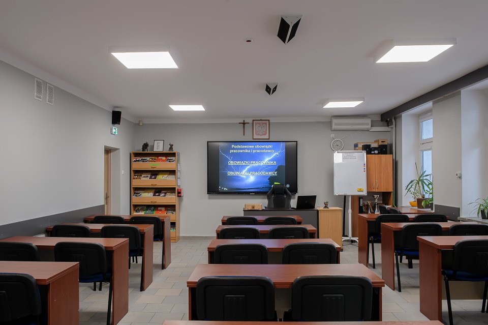 Sala lekcyjna z ekranem multimedialnym i rzędami stolików.