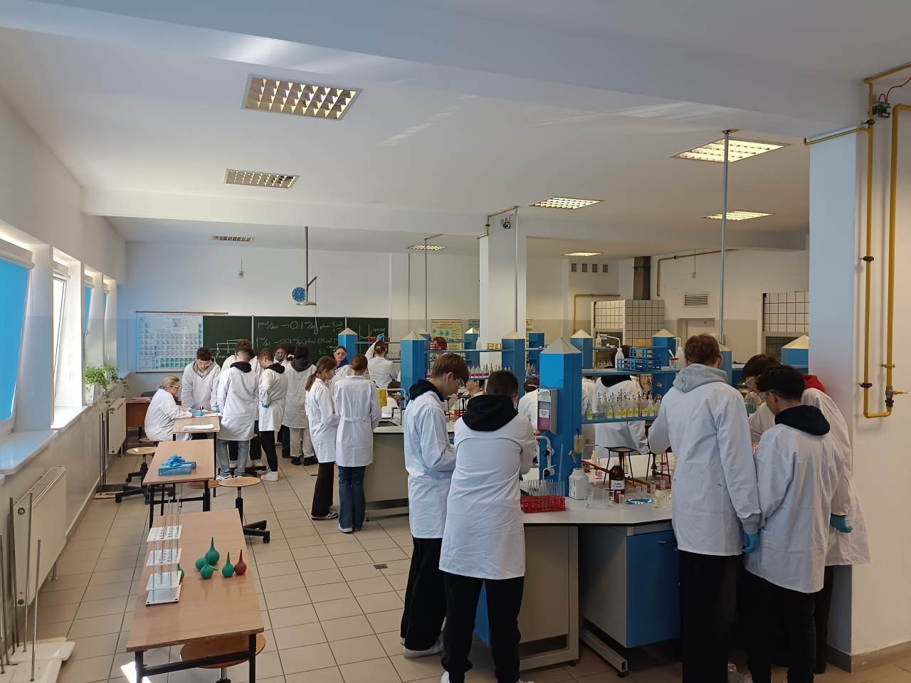 Uczniowie w fartuchach i rękawiczkach wykonują doświadczenia przy stanowiskach laboratoryjnych.