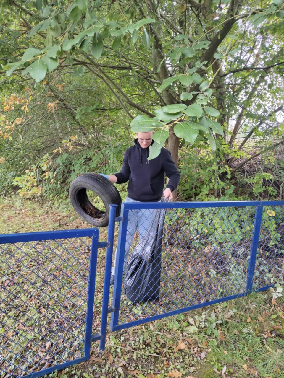 Uczestnik akcji podnosi znalezioną oponę przy zadrzewionym terenie.