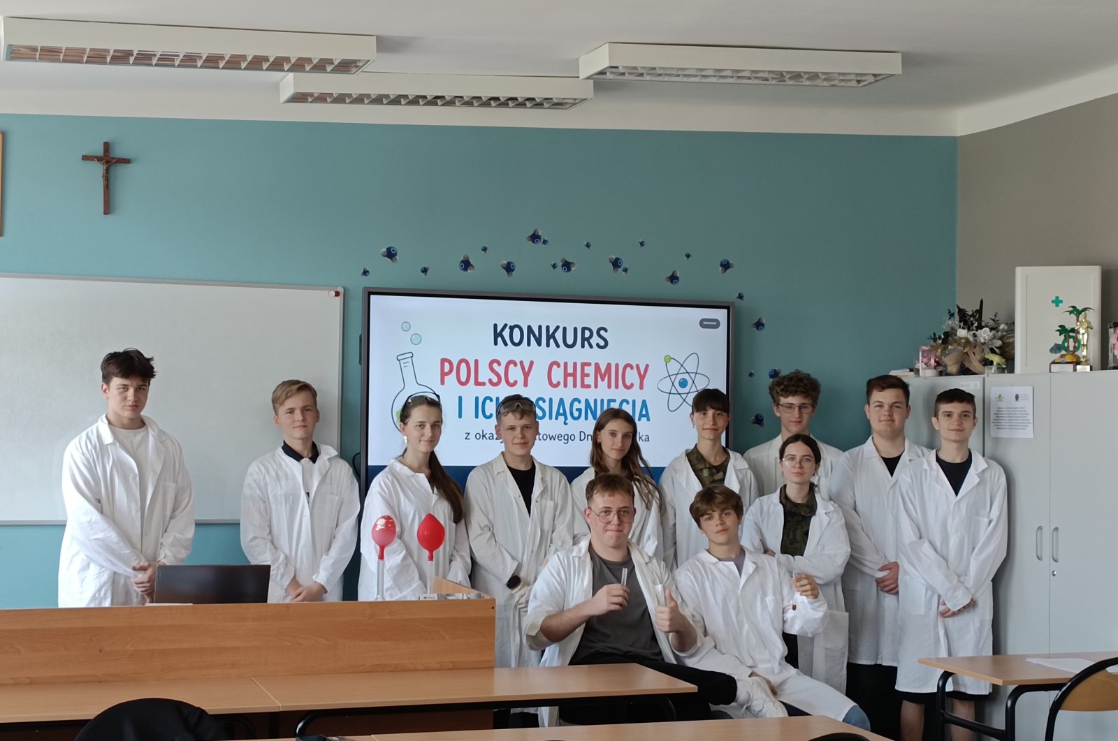 Uczestnicy Dnia Chemika w białych fartuchach, na tle prezentacji konkursu „Polscy chemicy i ich osiągnięcia”.