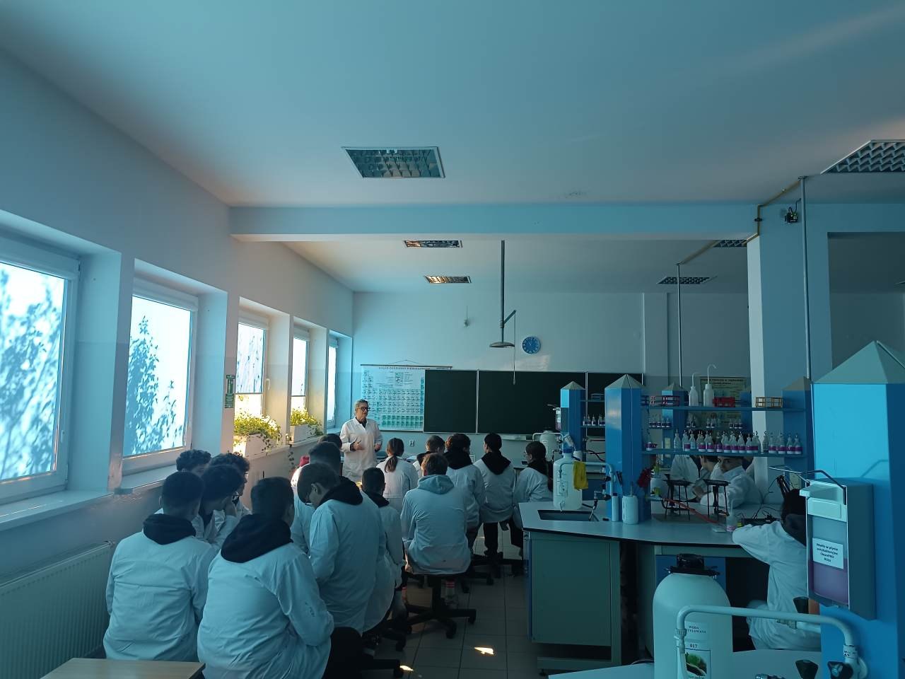 Prowadząca omawia zagadnienia chemiczne z uczniami siedzącymi przy stanowiskach laboratoryjnych.