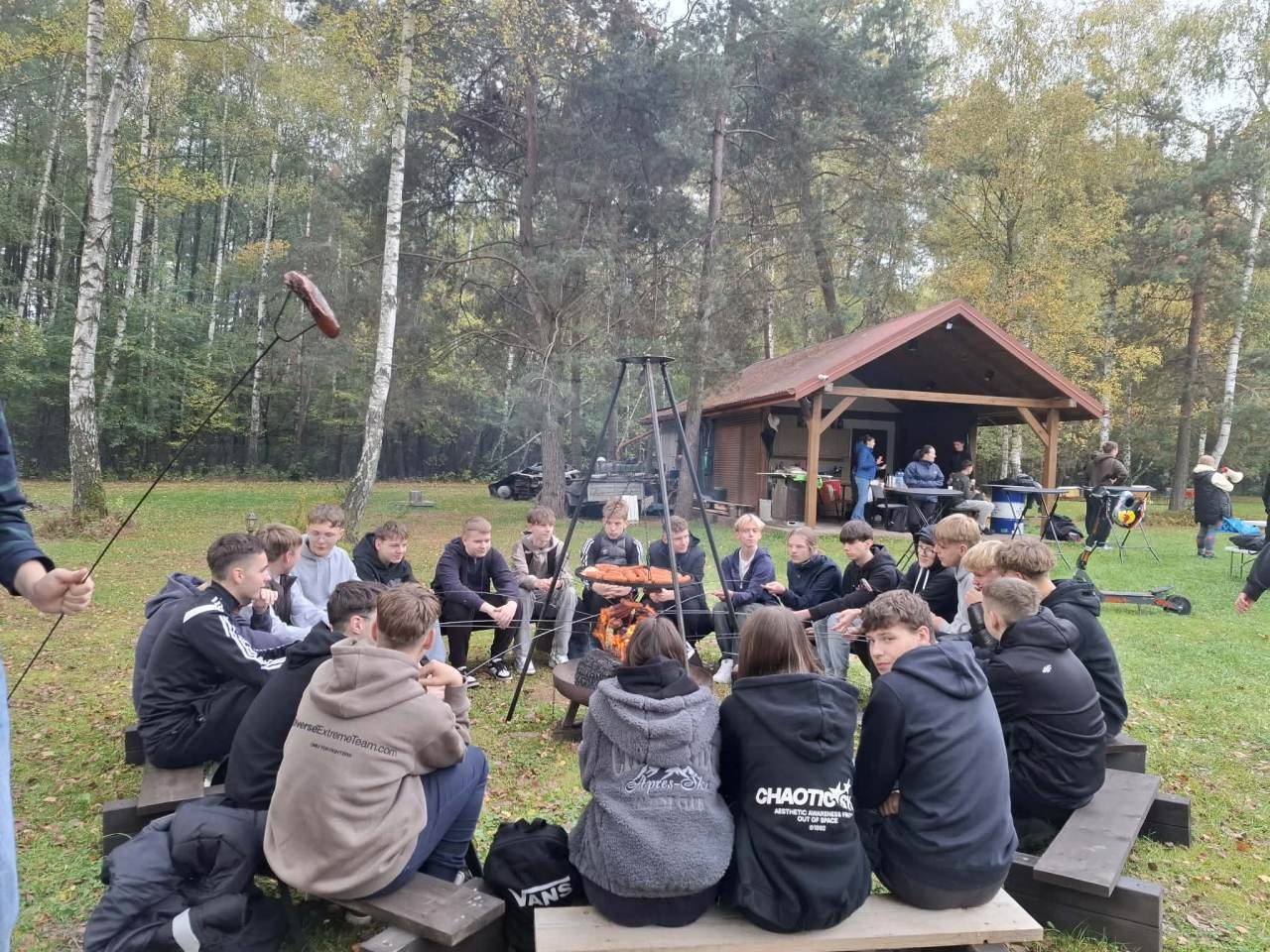 Uczestnicy wycieczki integracyjnej siedzą wokół ogniska i pieką kiełbaski.