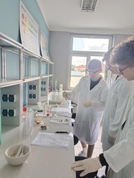 Uczniowie w fartuchach i rękawicach pracują przy laboratoryjnym blacie.