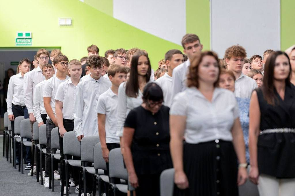 Uczestnicy uroczystości stoją w rzędach między krzesłami.