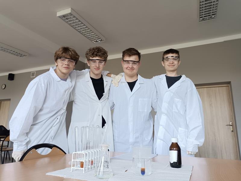 Czterech uczestników doświadczeń w fartuchach i okularach ochronnych przy stole laboratoryjnym.