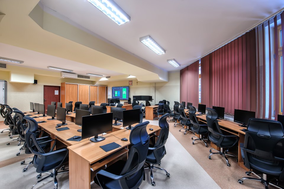 Pracownia komputerowa z fotelami i monitorami na drewnianych biurkach.