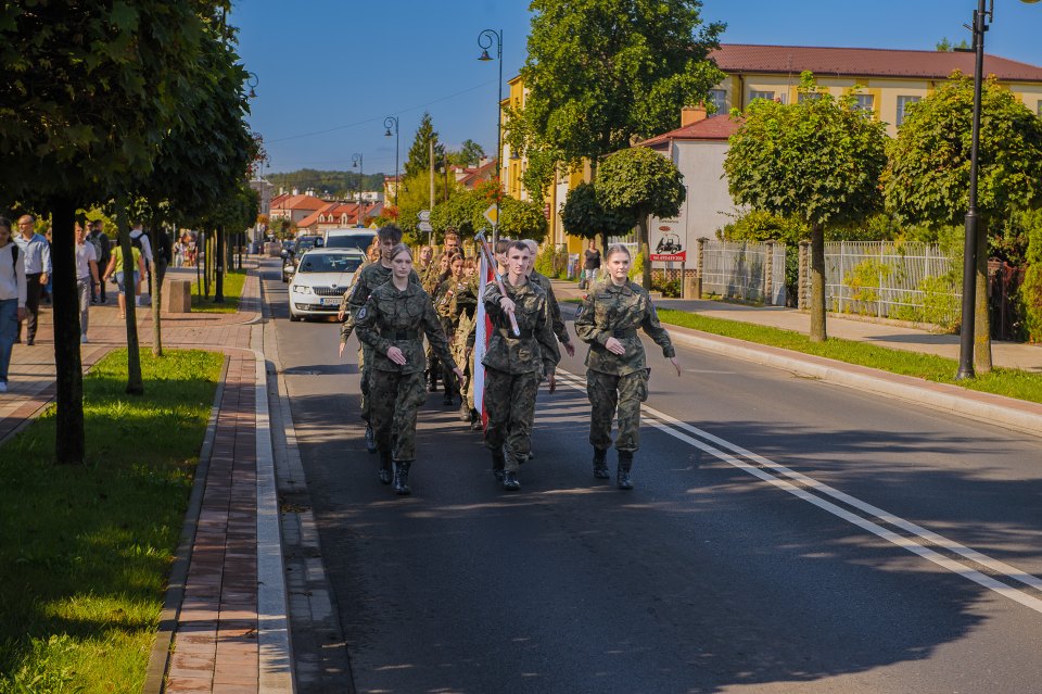 Poczet i osoby w mundurach maszerują ulicą podczas uroczystości.