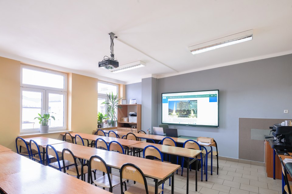 Sala lekcyjna z projektorem, ekranem i rzędami ławek.